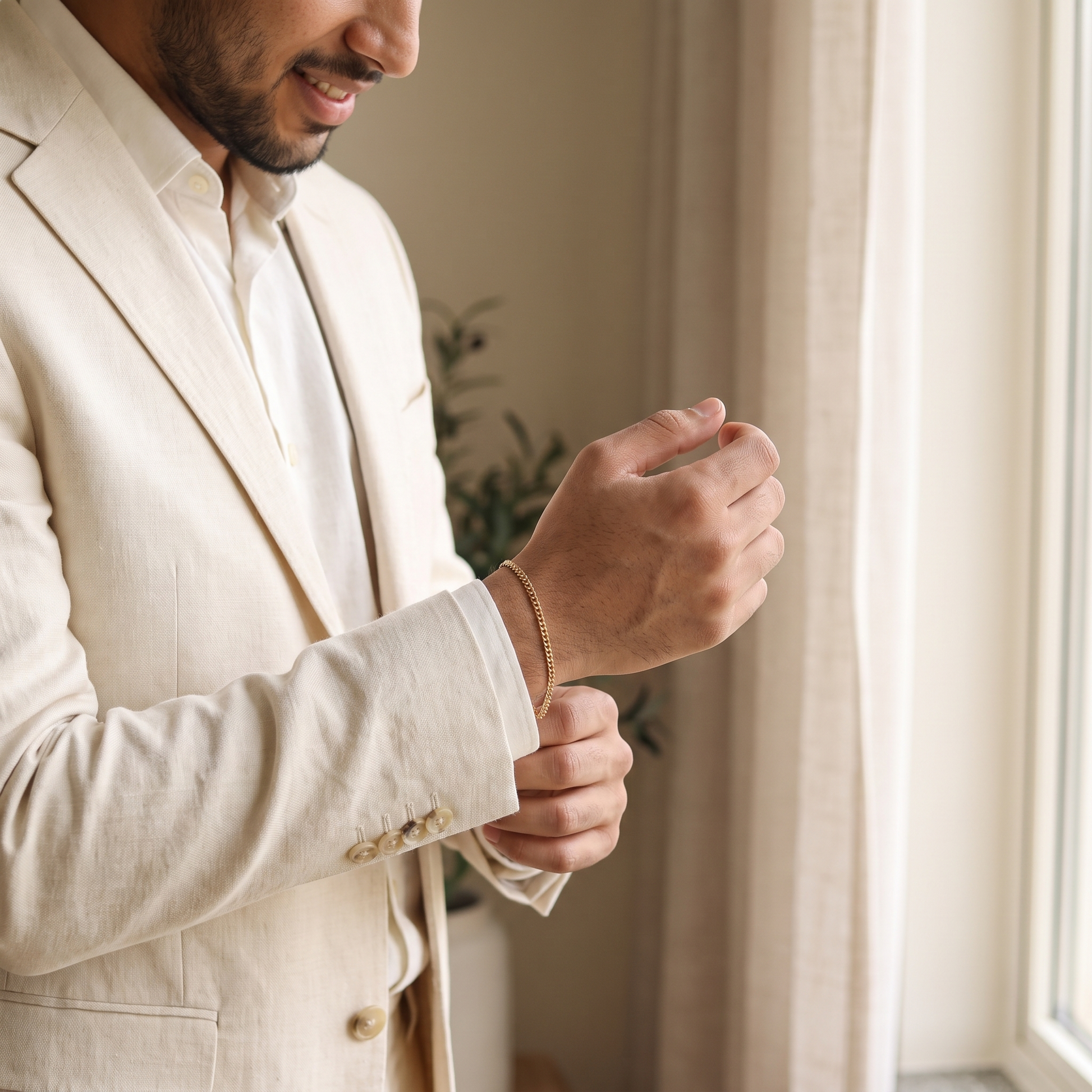 Bräutigam im Creme-Anzug mit zartem goldenem Cuban-Link-Armband beim Ankleiden am Fenster